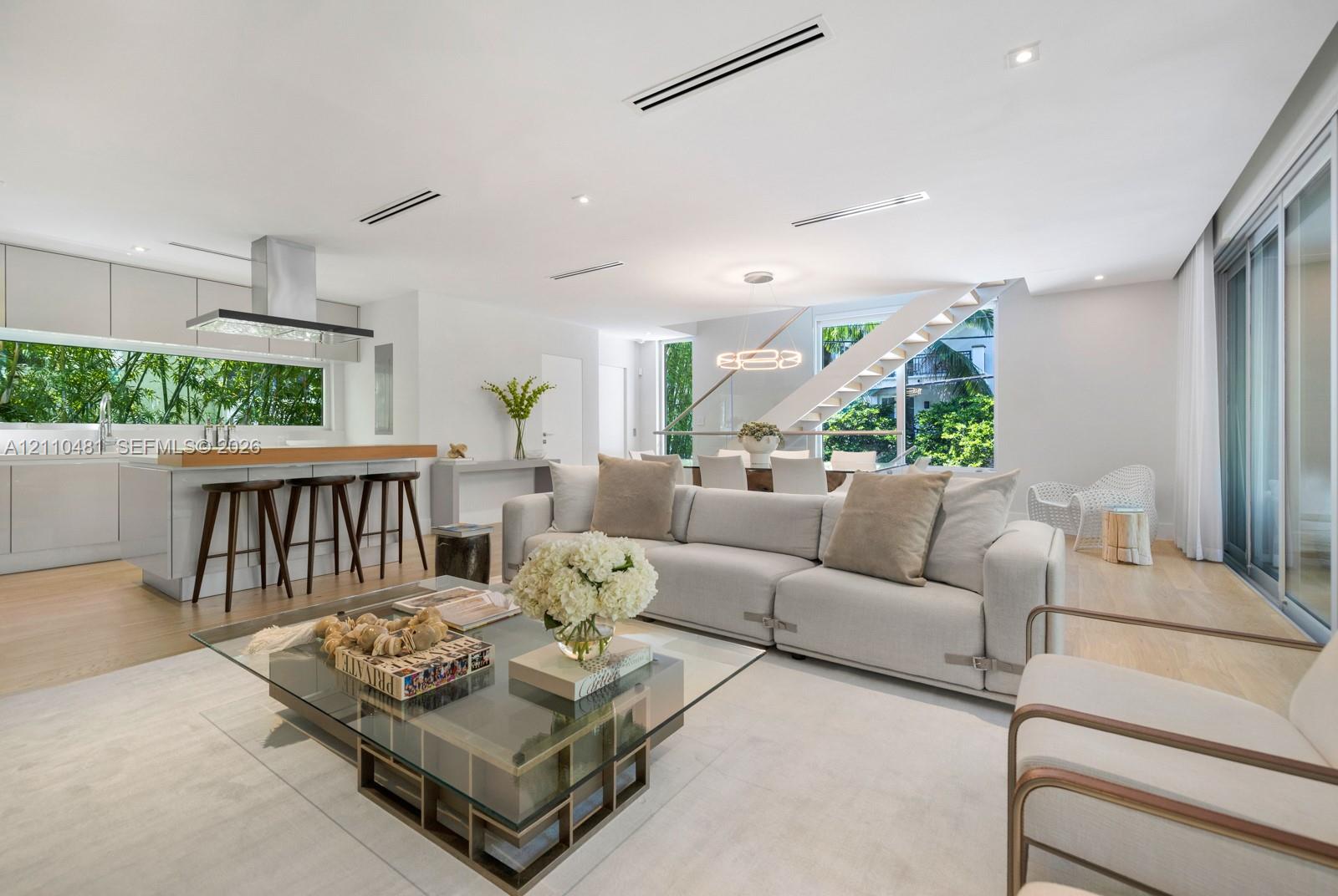 Open-concept interior featuring light wood-finish flooring, linear recessed lighting, a modern floating staircase with glass railings, large horizontal windows, and a stainless steel kitchen ventilation hood