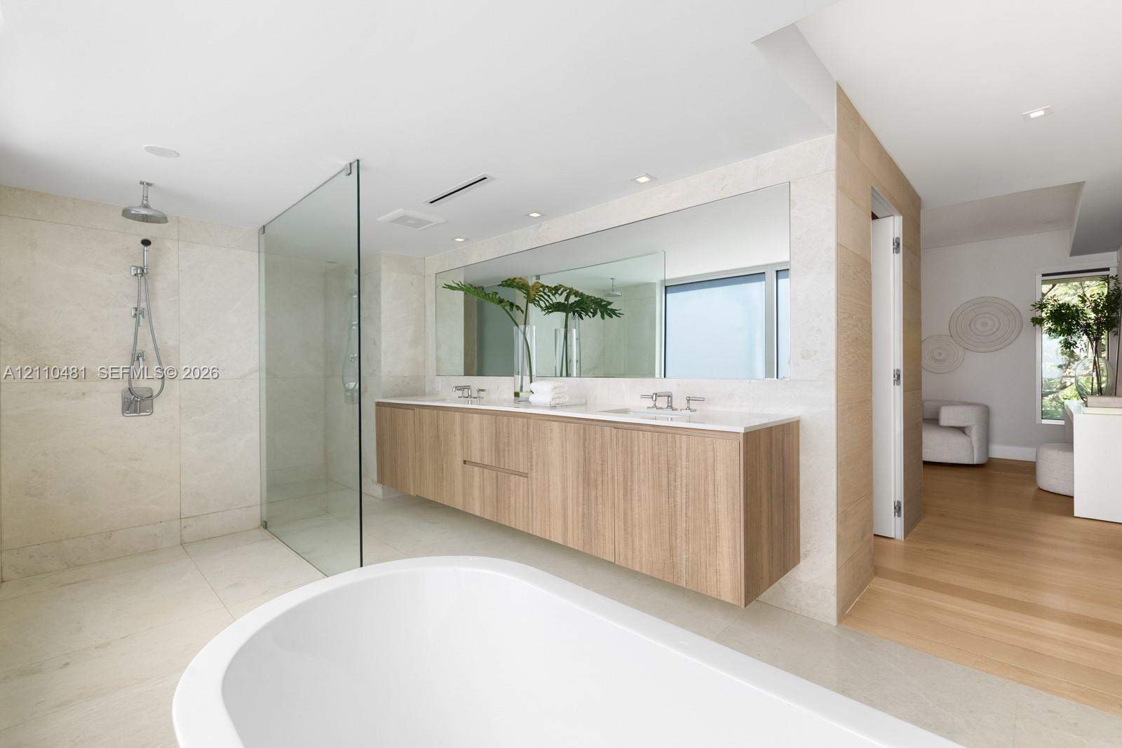 Floating wood-finish vanity with dual sinks, stone-tile shower with glass partition, freestanding soaking tub, large wall-to-wall mirror, and recessed lighting