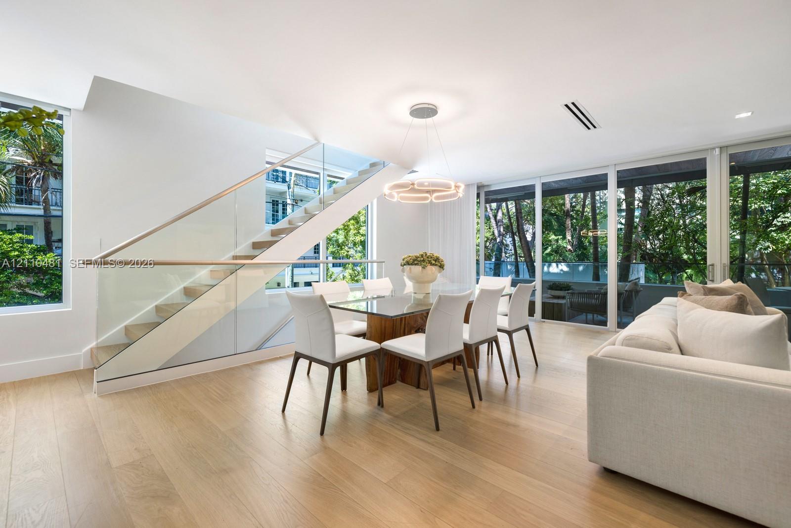 Open-plan interior featuring light wood-finish flooring, floor-to-ceiling glass panels, and a staircase with glass railings and a wood handrail
