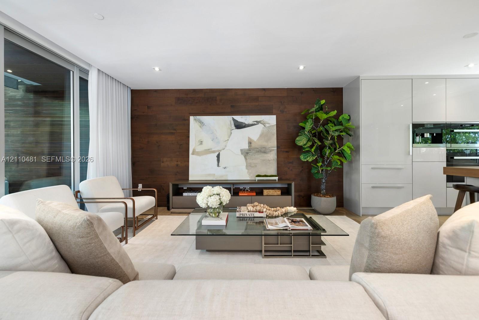 Open-concept interior featuring wood-paneled accent wall, recessed lighting, expansive floor-to-ceiling windows, and white cabinetry with integrated appliances