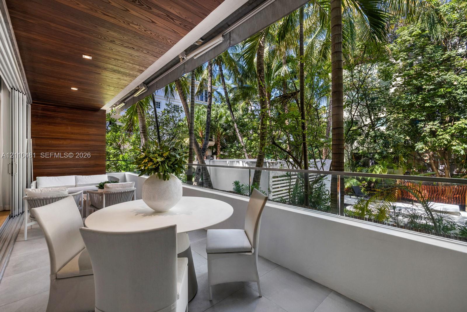 Covered balcony featuring wood-paneled ceiling, recessed lighting, tile flooring, and a glass-paneled railing overlooking mature palm trees