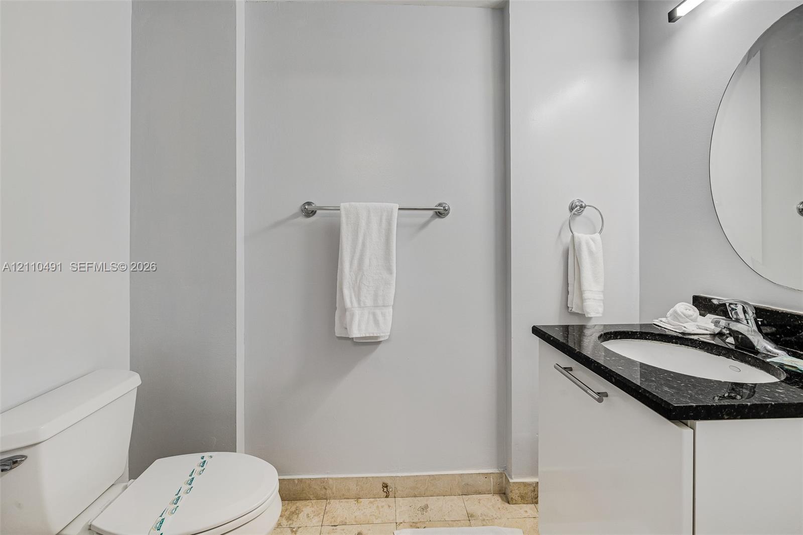 White vanity with integrated basin, dark stone-finish countertop, polished chrome faucet, round wall-mounted mirror, and tile flooring