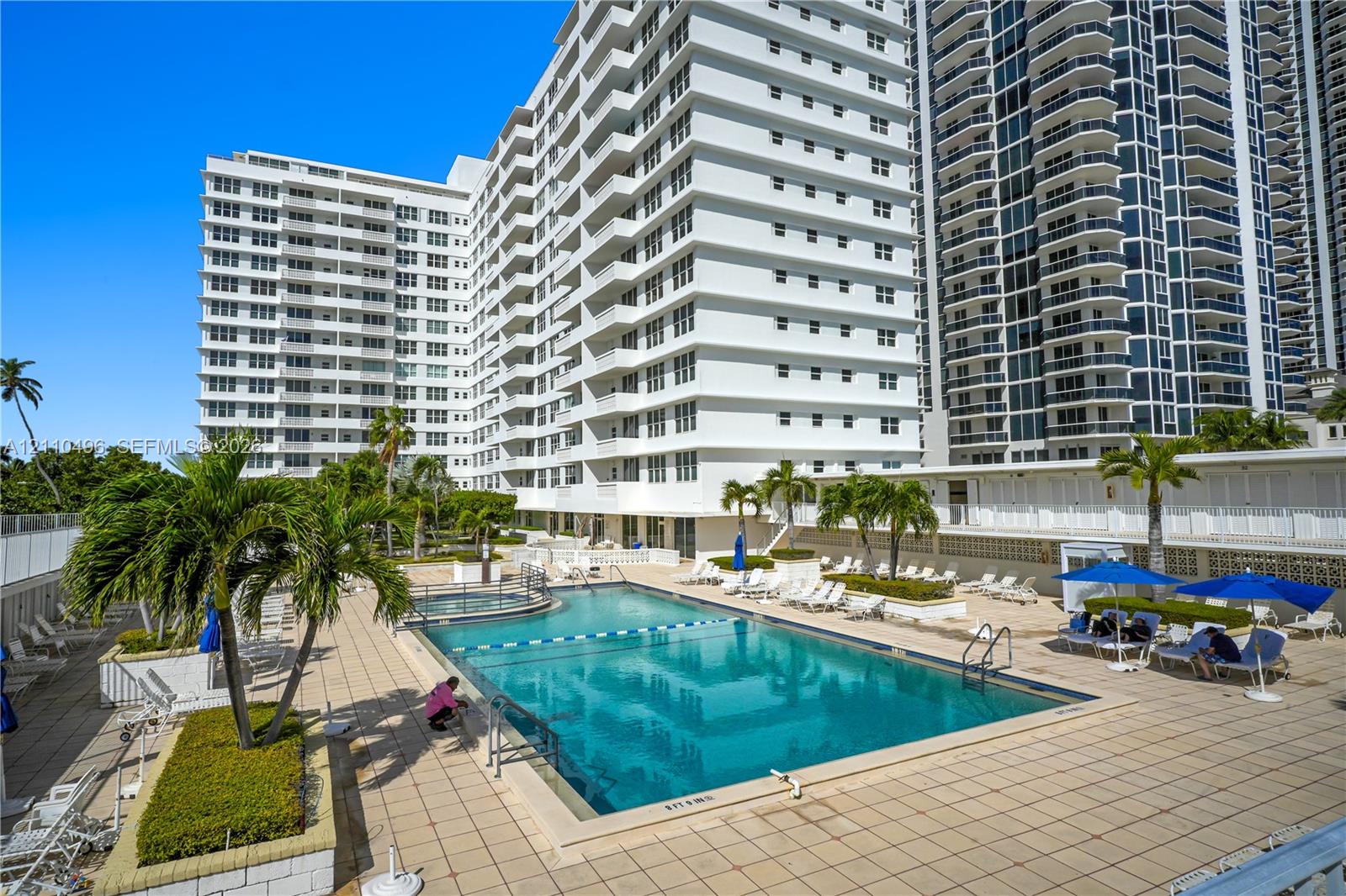 High-rise residential exterior featuring stacked private balconies, ground-level swimming pool, integrated spa, tiled pool deck, and mature palm landscaping