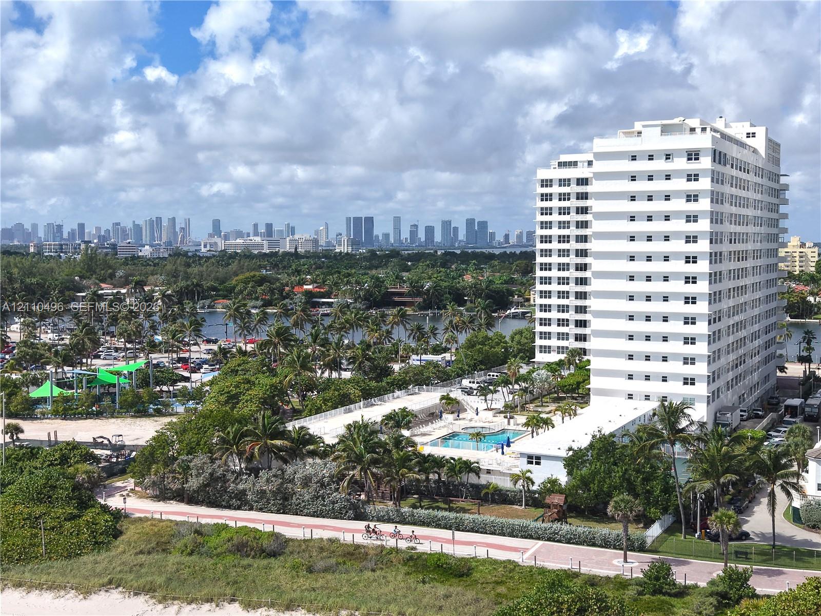 High-rise residential building featuring an outdoor swimming pool, paved pedestrian walkway, and mature palm landscaping with a backdrop of a coastal skyline