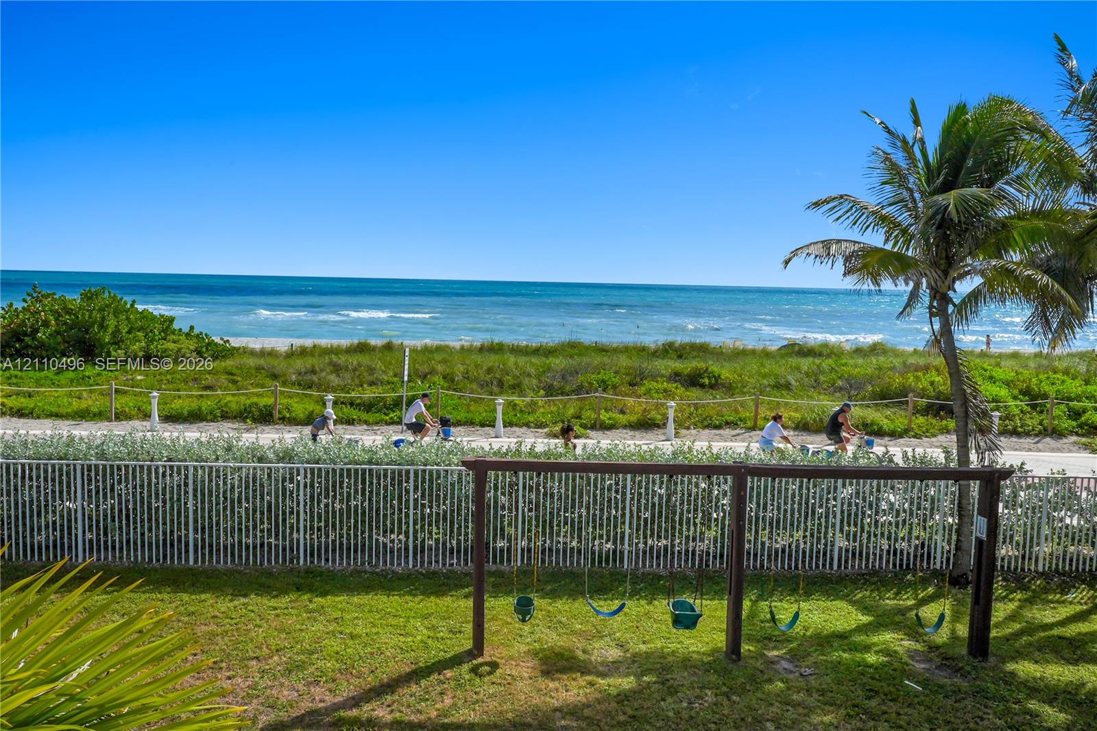 Direct oceanfront property featuring a grassy lawn, a wooden swing set, and a perimeter fence with direct access to a paved coastal walkway