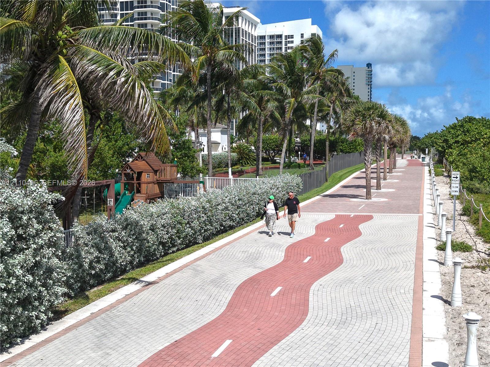 Paved multi-use path featuring decorative interlocking brick pavers, mature palm trees, and native landscape screening