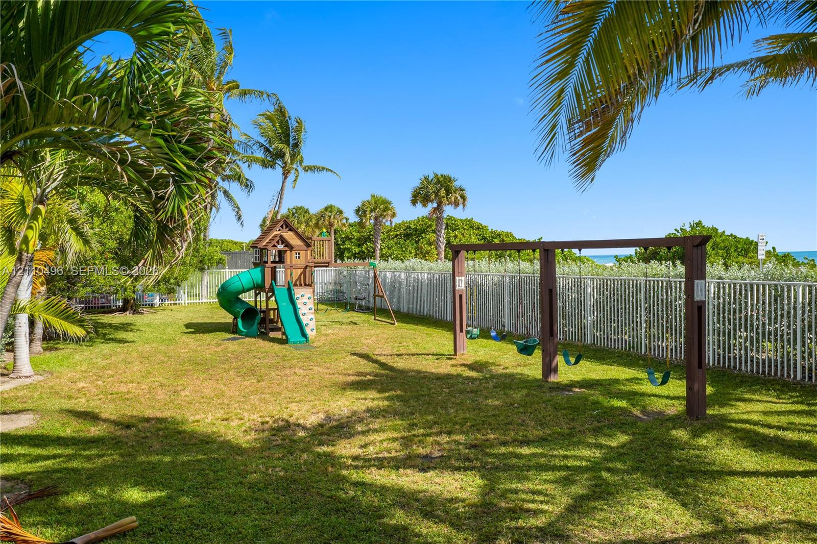 Backyard featuring a grass lawn, mature palm trees, perimeter fencing, and a built-in wooden play structure with a slide and swing set