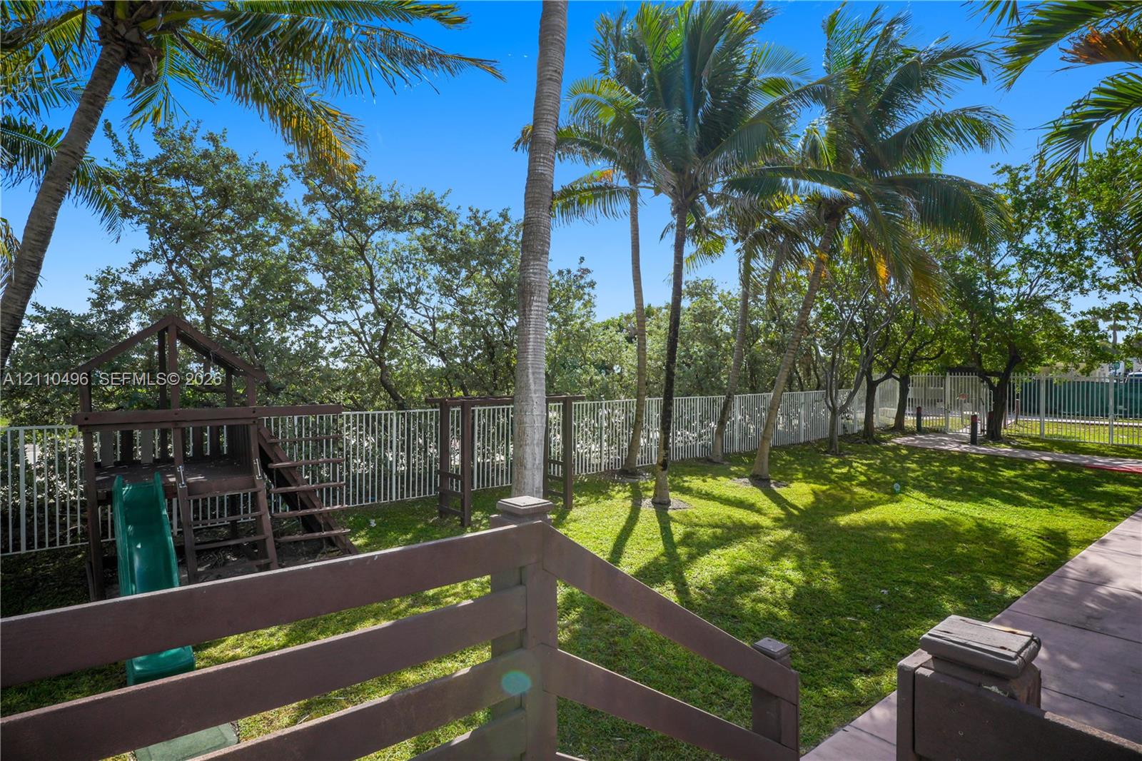 Rear yard featuring a manicured lawn, established palm trees, perimeter metal fencing, and wood-finish decking