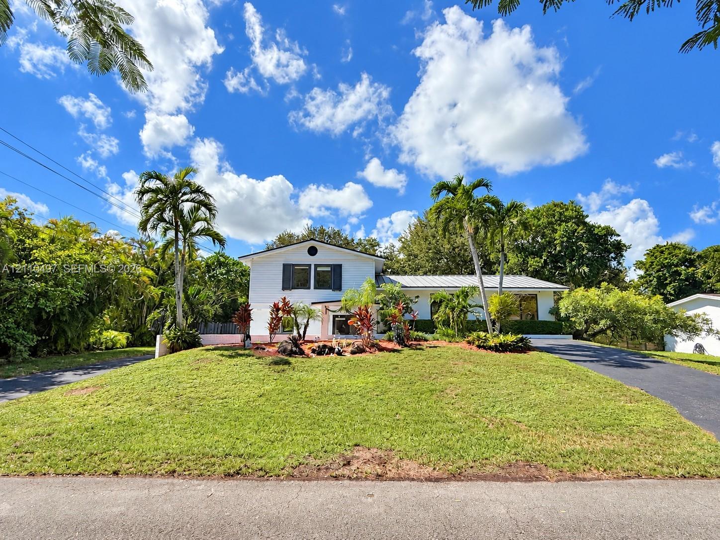 Exterior facade featuring white siding, dark shutters, a metal roof, mature palm trees, and an asphalt driveway