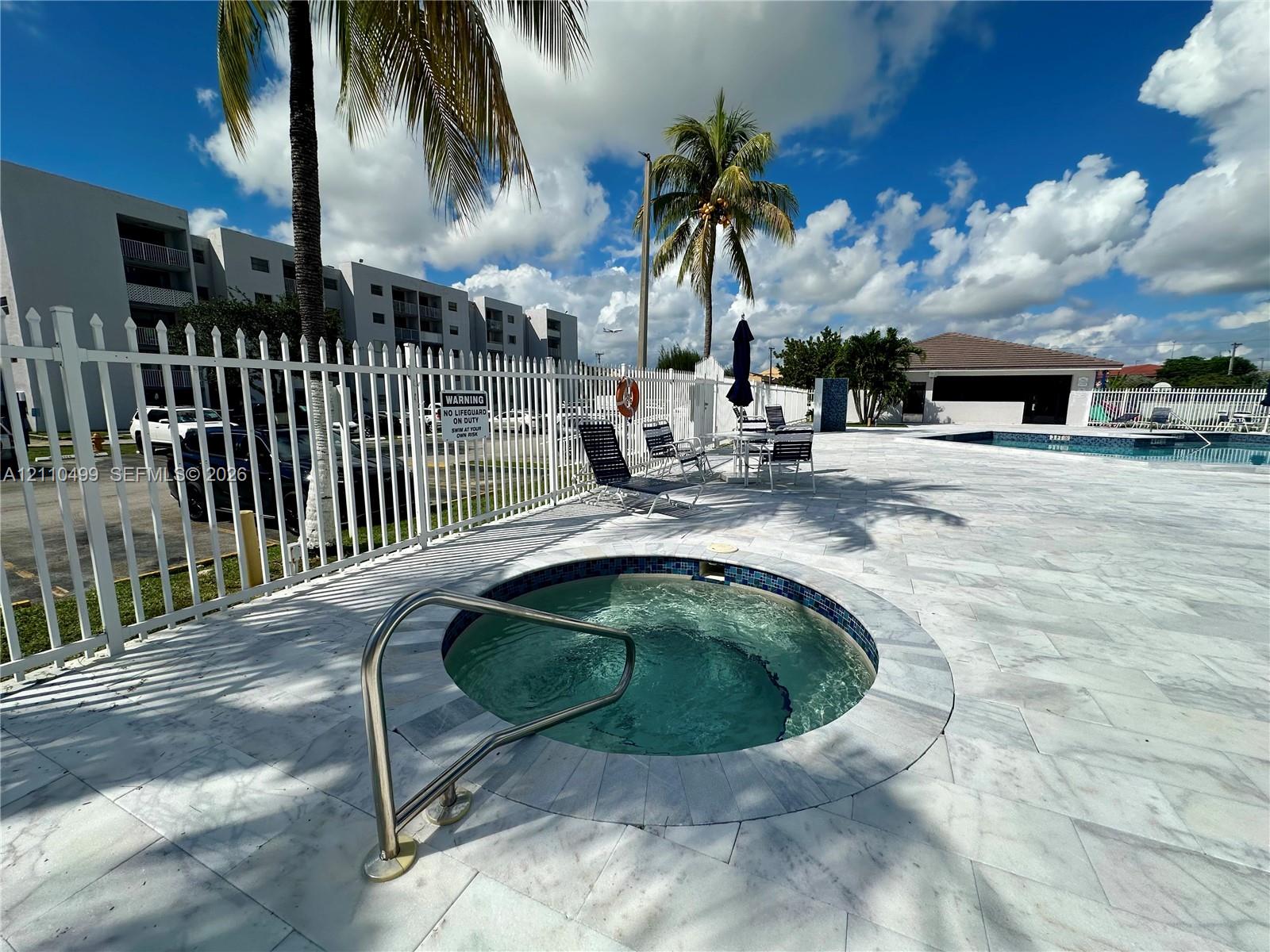 In-ground spa with a stainless steel handrail, surrounded by light-toned stone decking and enclosed by white metal fencing