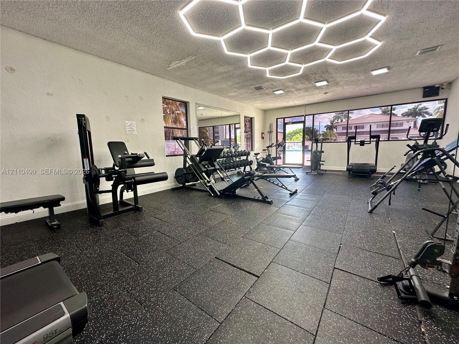 Fitness room featuring rubberized tile flooring, honeycomb-pattern ceiling lighting, floor-to-ceiling windows, and wall-mounted mirror panels