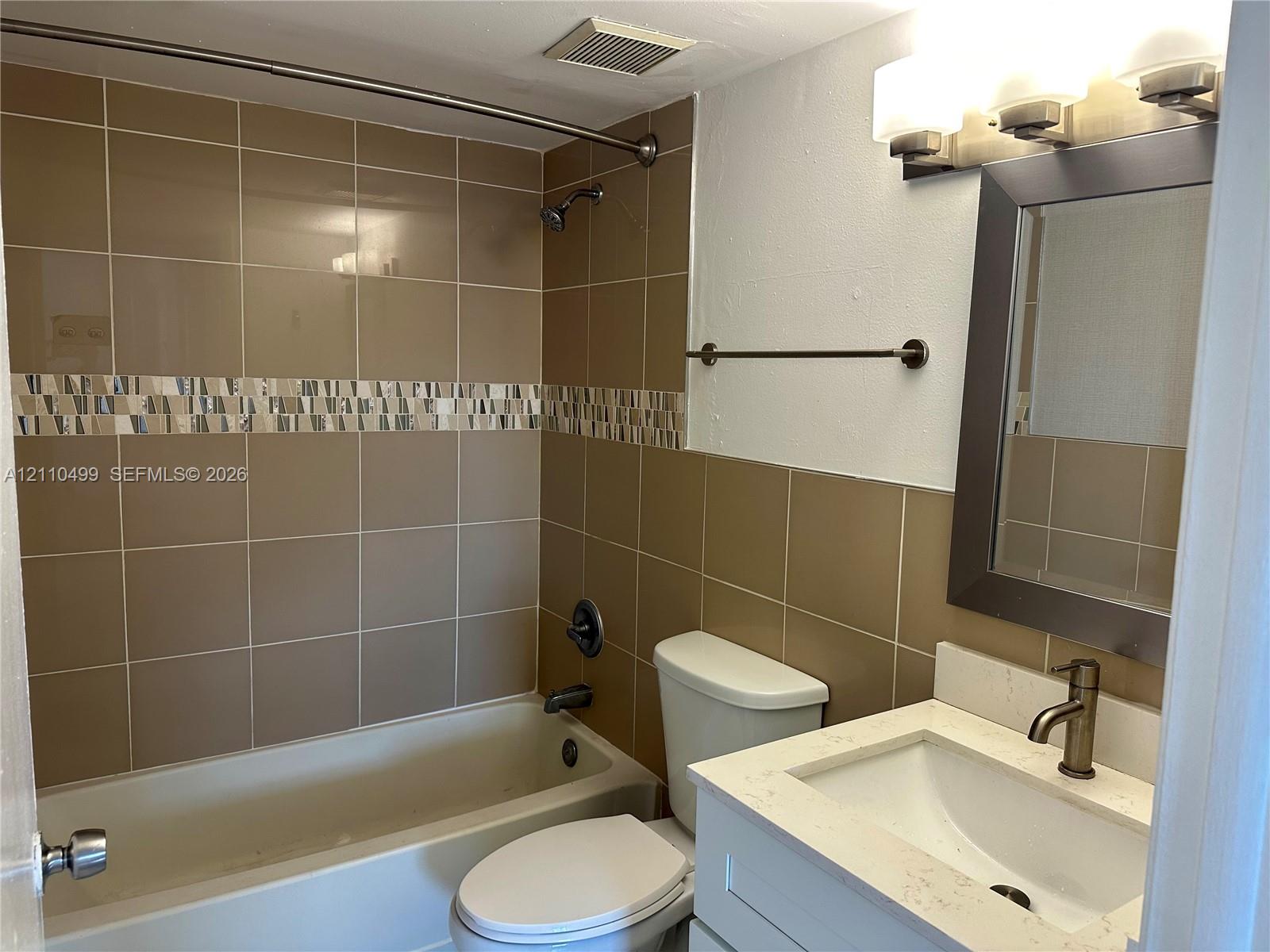 Bathroom featuring a tub and shower combination with tile surround and decorative accent border, white vanity with stone-finish countertop, undermount sink, and wall-mounted vanity light