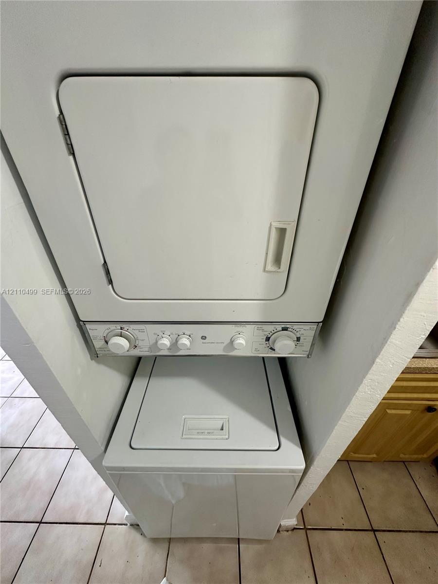 Stacked laundry unit with top-loading washer and front-loading dryer, set within an alcove featuring ceramic tile flooring