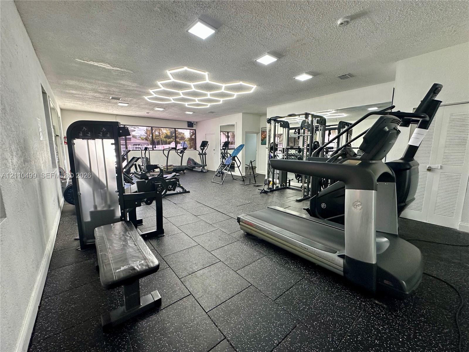 Fitness center featuring durable rubberized flooring, large-scale wall-mounted mirrors, recessed LED lighting, and a prominent hexagonal ceiling light fixture
