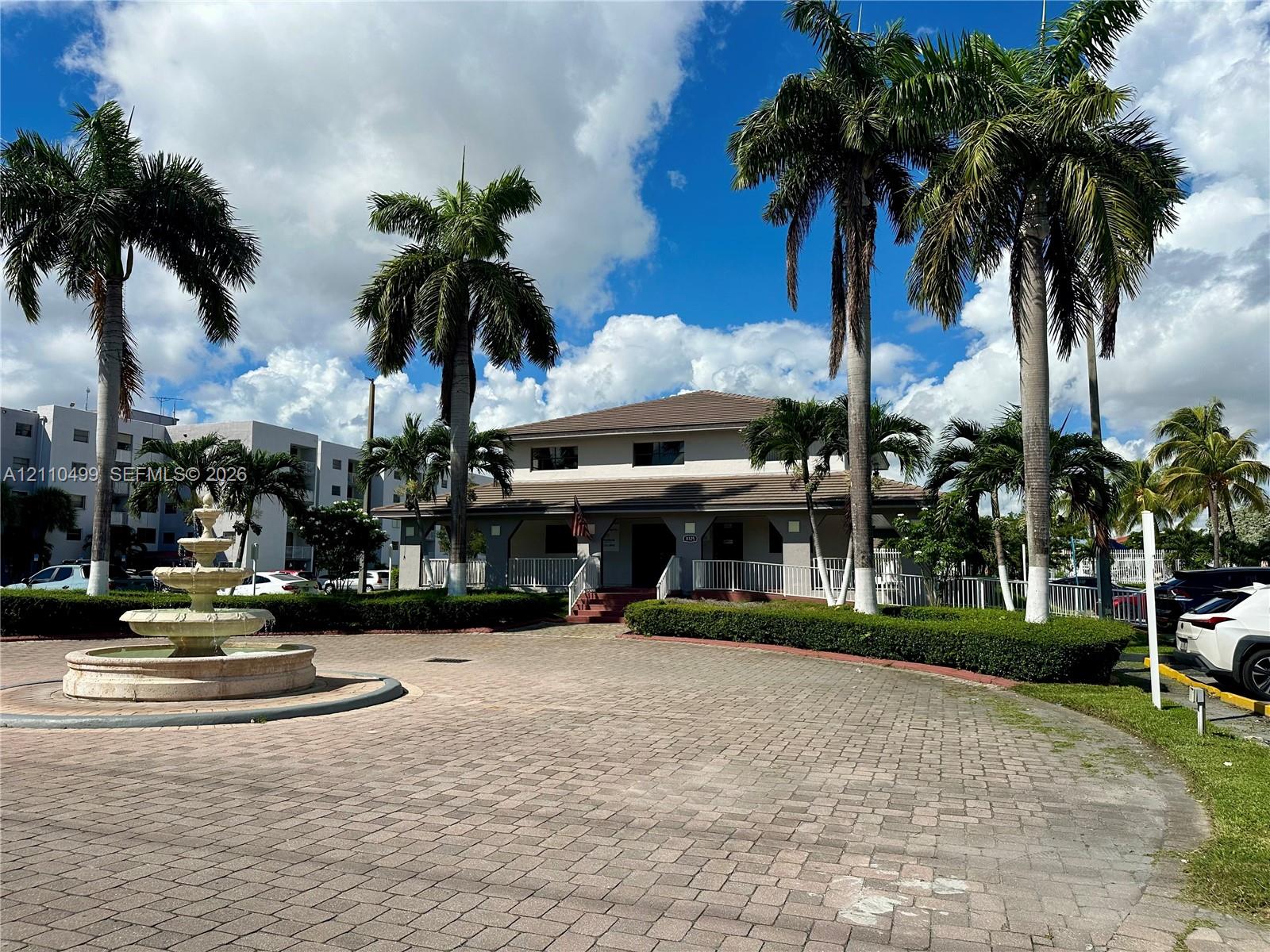 Paver brick circular driveway featuring a tiered stone fountain, manicured hedges, and mature palm trees