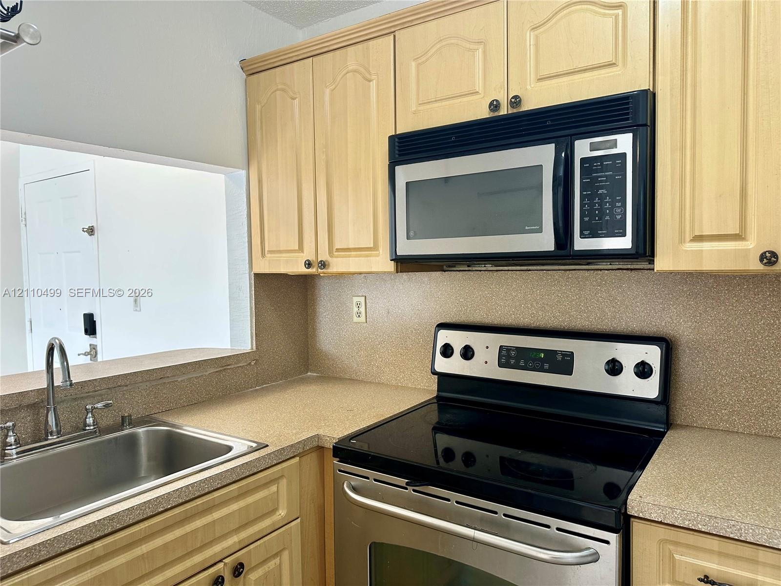 Kitchen featuring light-finish cabinetry, stainless steel microwave, stainless steel range with electric glass cooktop, solid-surface countertops, and a stainless steel sink with a high-arch faucet Kitchen featuring light-finish cabinetry, stainless steel microwave, stainless steel range with electric glass cooktop, solid-surface countertops, and a stainless steel sink with a high-arch faucet