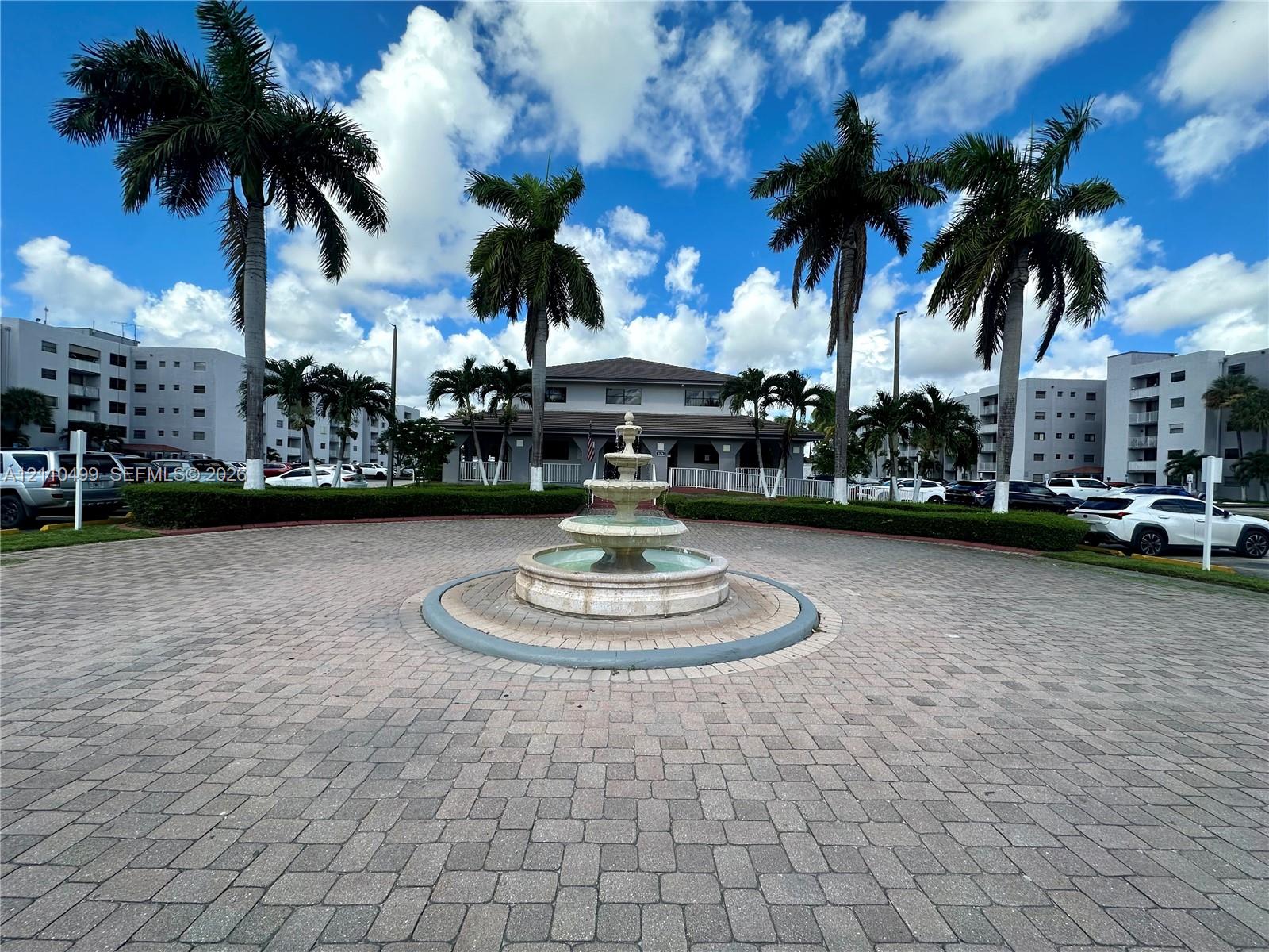 Circular paver driveway featuring a tiered stone fountain, mature palm trees, and manicured hedges