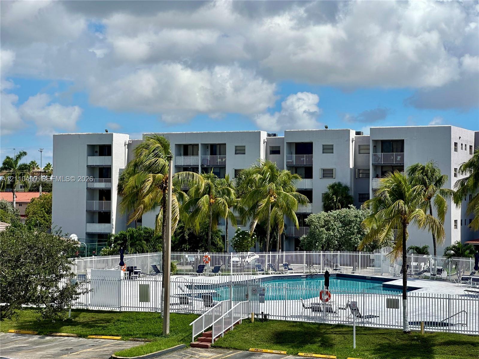 Multi-story residential building featuring private balconies, swimming pool with perimeter fencing, and mature palm trees