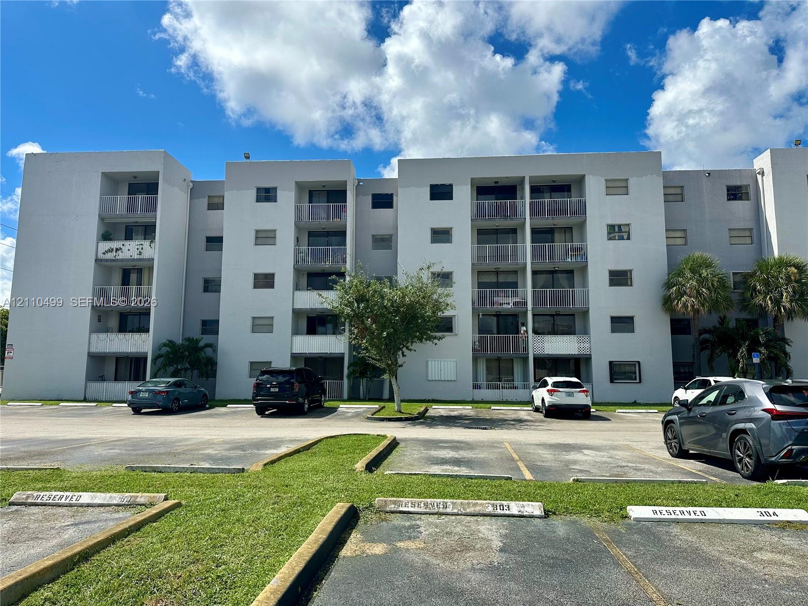 Mid-rise building exterior featuring a multi-story facade, private balconies with metal railings, and a paved surface parking lot
