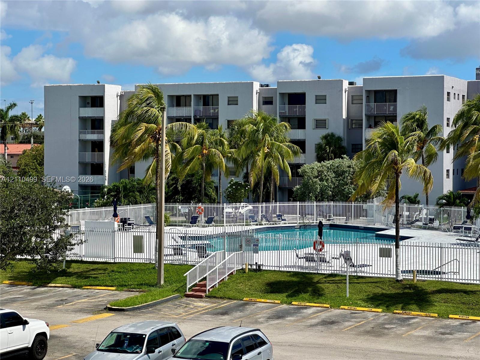 Multi-story residential building featuring private balconies, an enclosed swimming pool, metal perimeter fencing, and mature palm trees