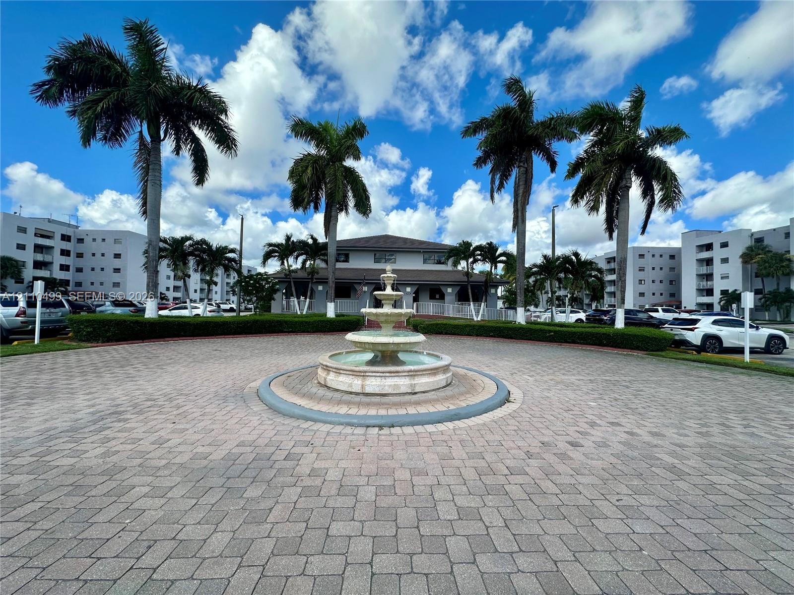 Circular paved motor court featuring a multi-tiered stone fountain, manicured hedges, and mature palm trees