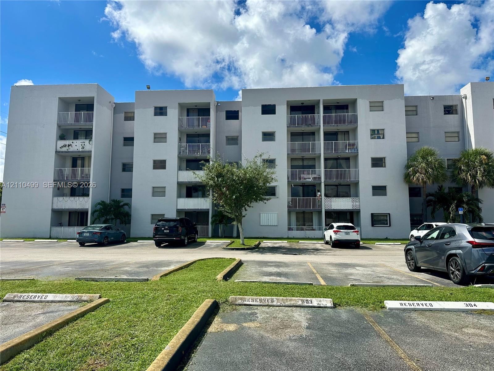 Multi-story concrete residential building featuring private balconies, designated parking spaces, and established palms