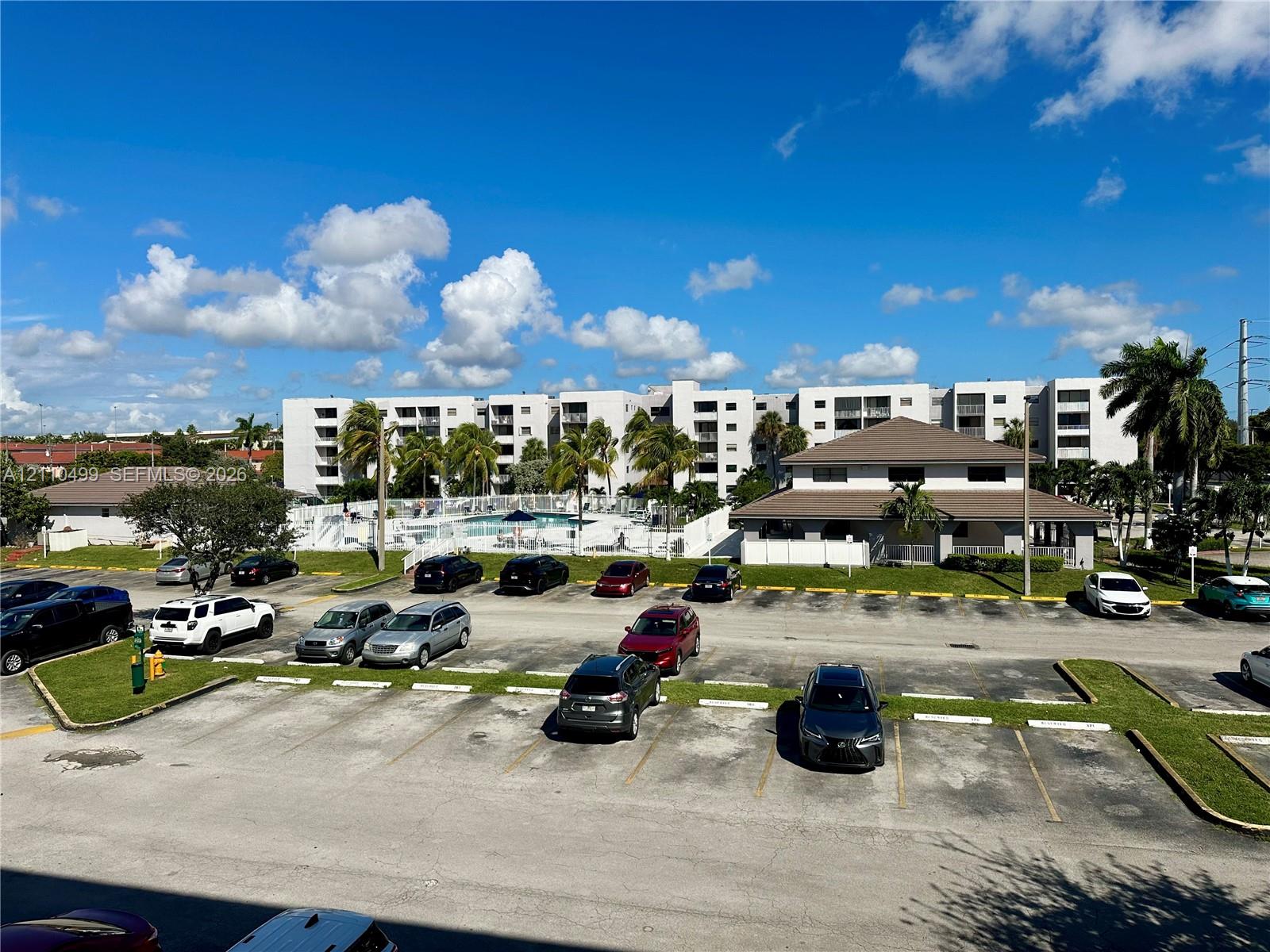 Multi-story residential complex featuring a swimming pool, mature palm trees, paved parking lot, and ground-level common area building