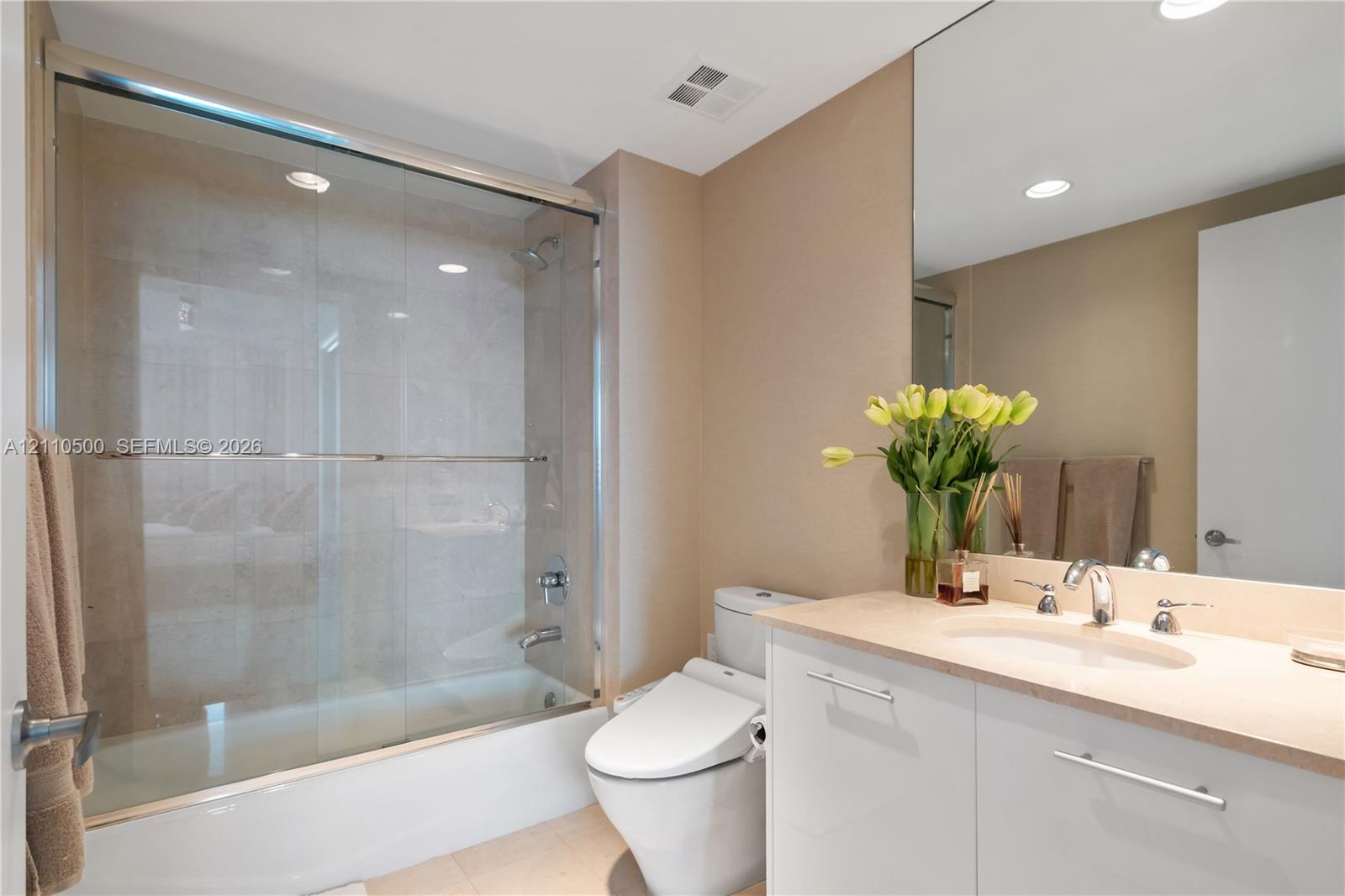 Bathroom featuring a tub-shower combination with glass enclosure, white vanity with stone-surfaced countertop, integrated oval basin, and a large wall-mounted mirror