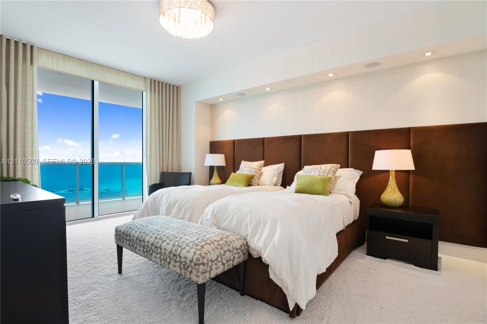 Floor-to-ceiling glass sliding doors opening to a balcony, custom brown upholstered headboard wall, recessed ceiling lighting, and a drum-style chandelier