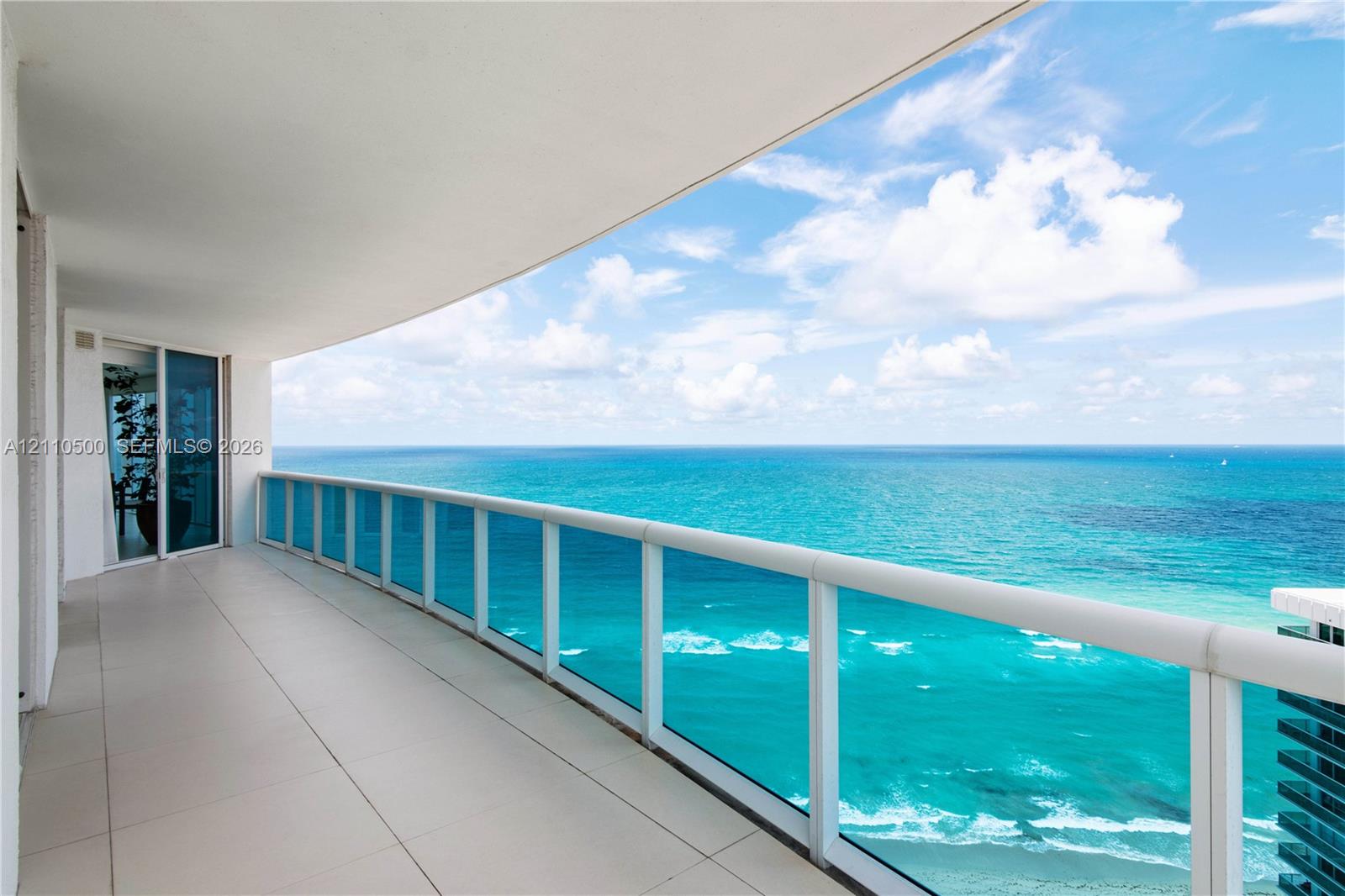 Curved wrap-around balcony with oversized tile flooring, full-height glass railings, and expansive ocean frontage