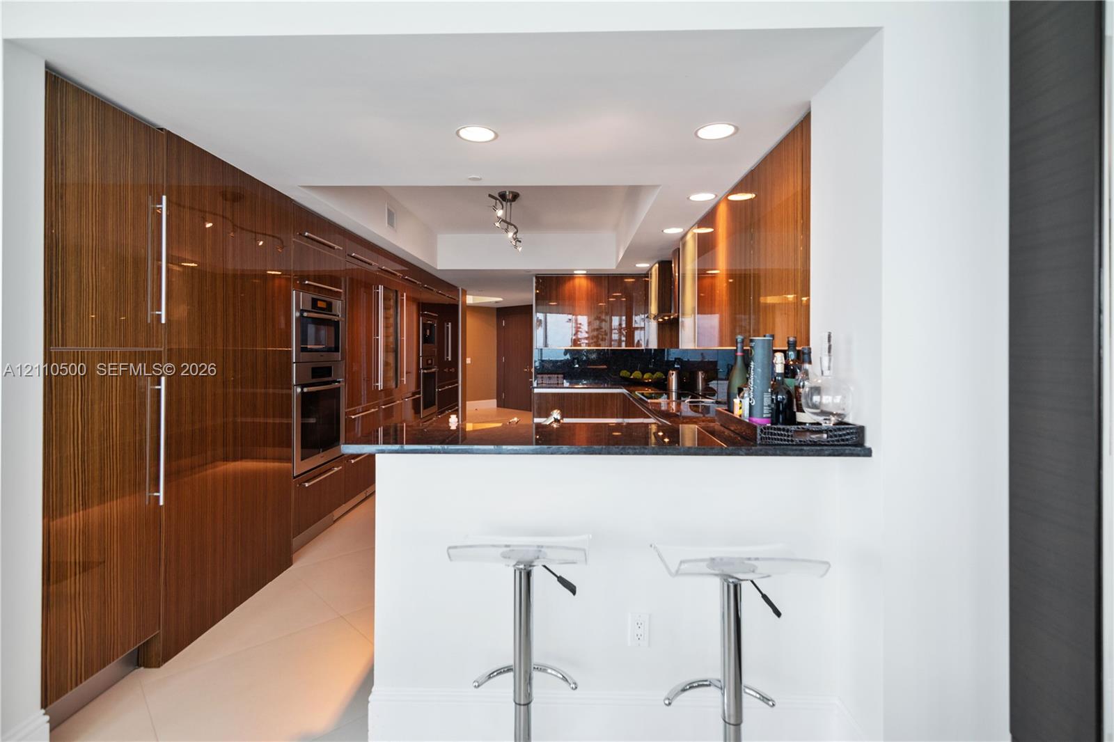 High-gloss wood-finish cabinetry, built-in stainless steel double wall ovens, dark stone countertop, and recessed ceiling lighting