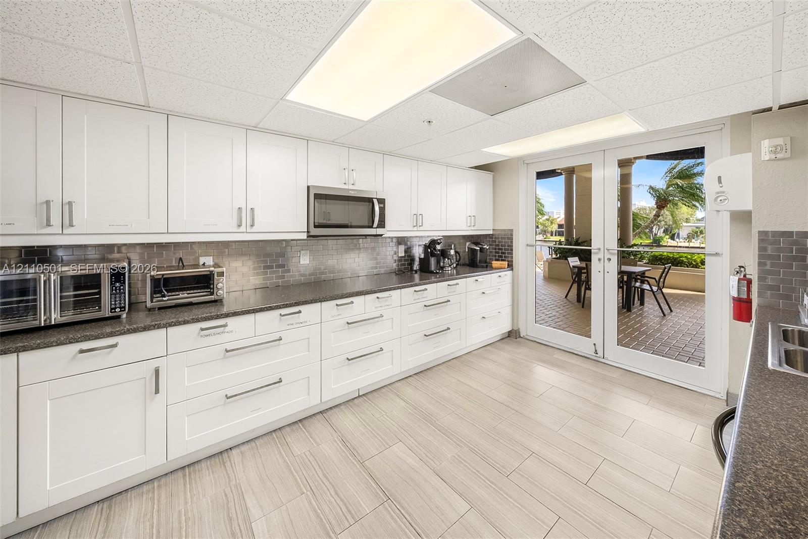 Kitchen featuring white shaker-style cabinetry, dark stone-finish countertops, subway tile backsplash, light-toned tile flooring, and double French doors opening to an outdoor patio