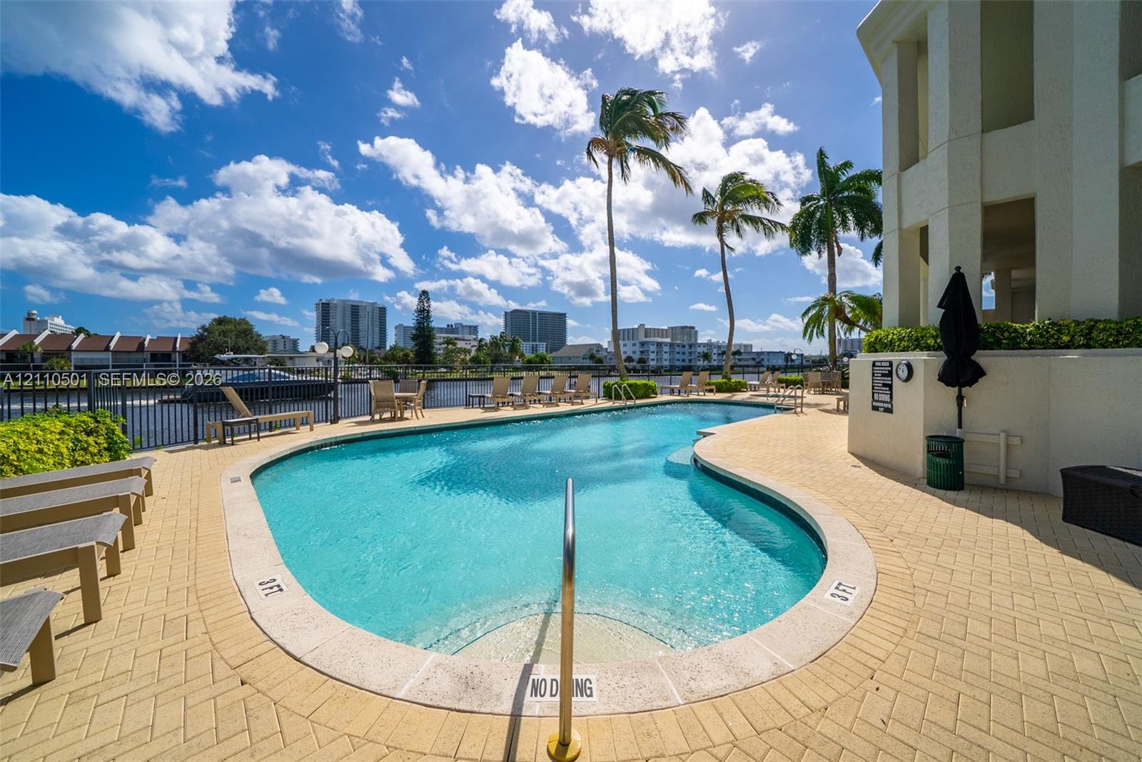 Waterfront swimming pool with paver decking and integrated stair entry, bordered by mature palm trees and metal perimeter fencing