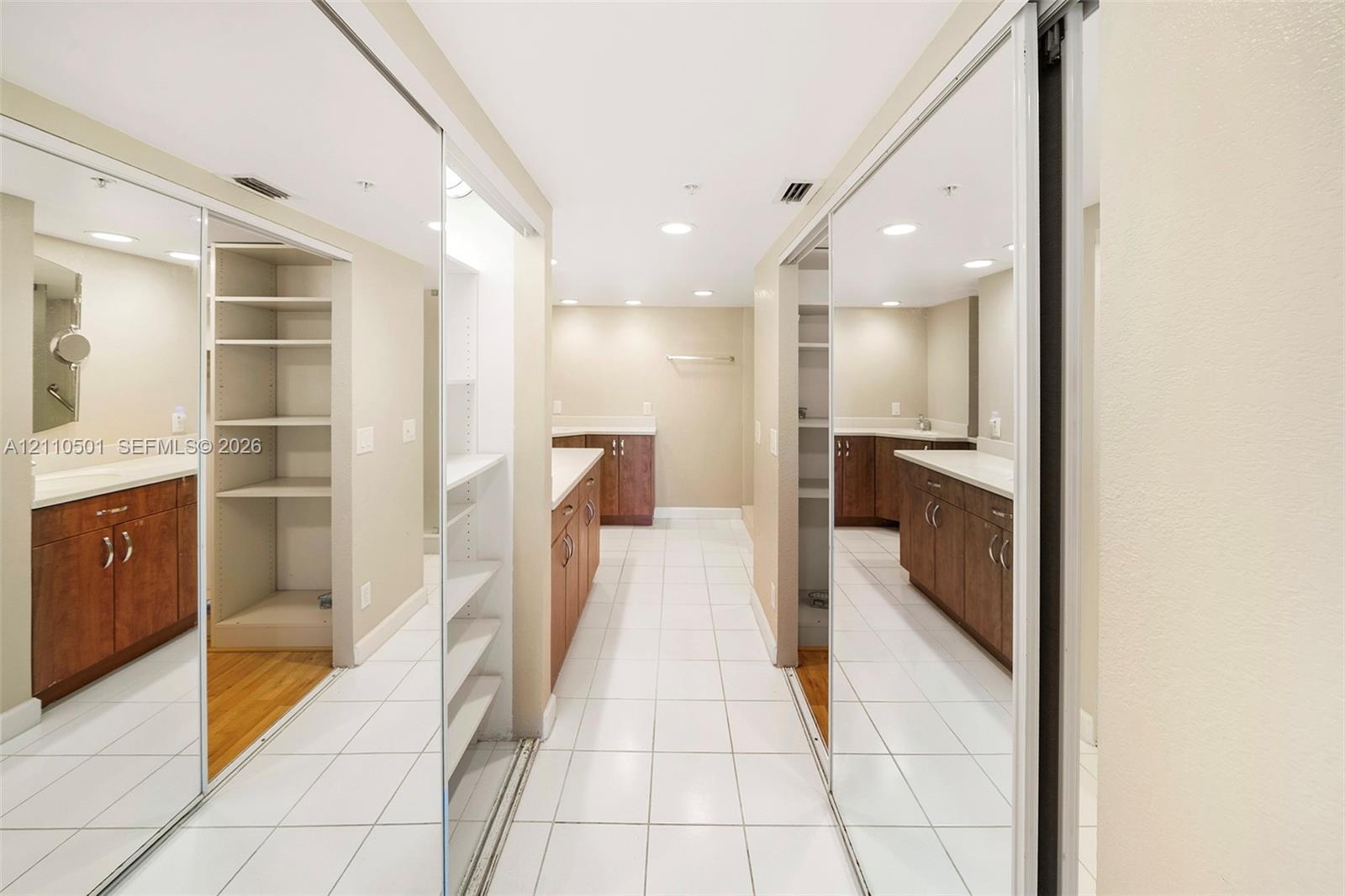 Walk-in dressing area featuring floor-to-ceiling mirrored sliding closet doors, built-in shelving, wood-finish cabinetry, neutral countertops, and white tile flooring