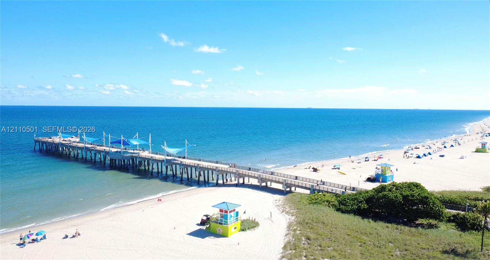 Coastal location featuring an extended ocean pier with structured shade canopies, direct sandy beach access, and surrounding natural vegetation