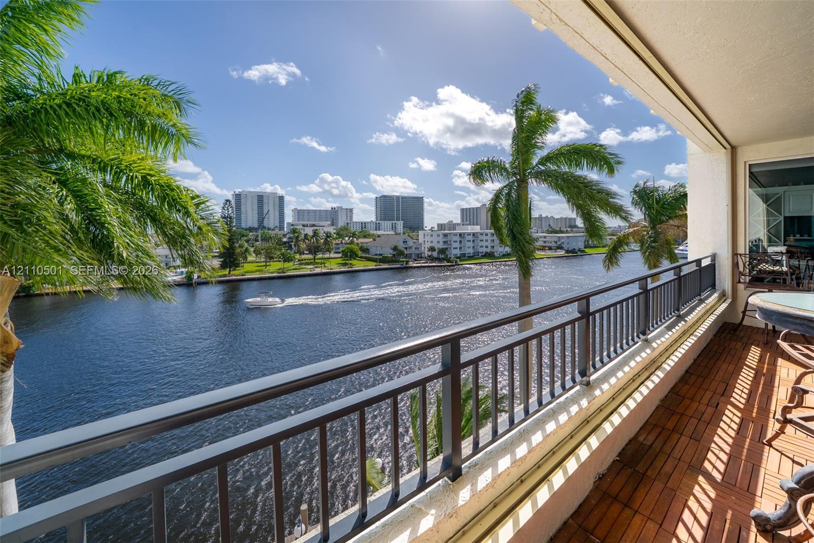 Covered balcony featuring wood-finish decking and a dark metal railing, overlooking a waterway with urban architecture in the background