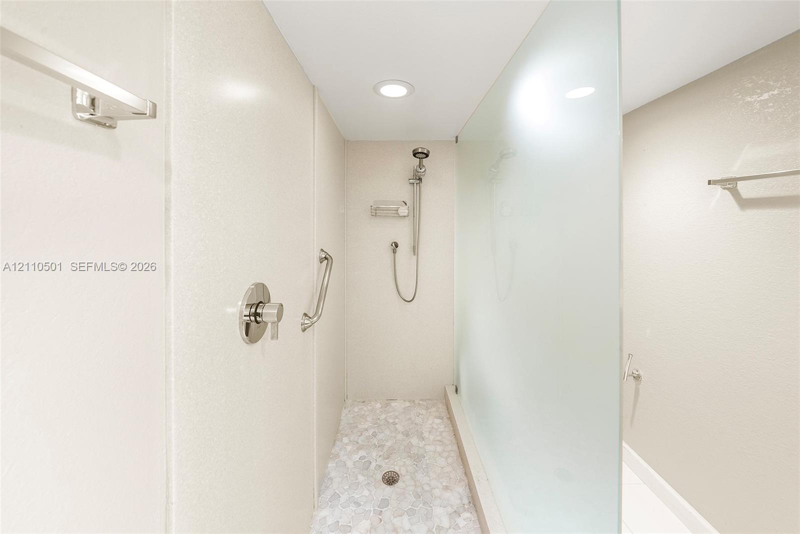 Walk-in shower featuring a frosted glass panel, pebble-stone flooring, brushed-nickel handheld shower head, and wall-mounted metal support bars