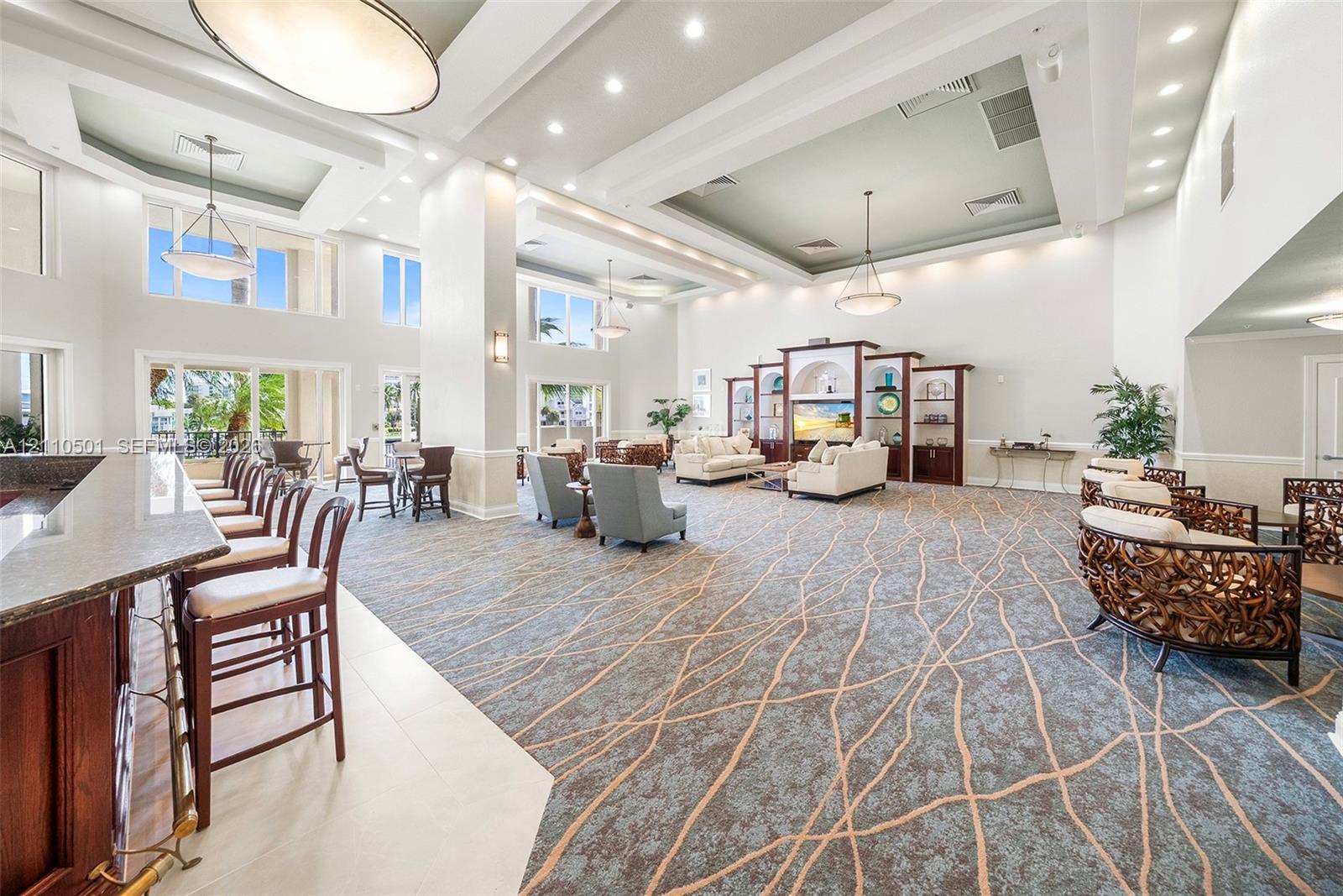 Expansive communal space featuring coffered ceilings, recessed lighting, integrated wooden shelving, a stone-topped bar with built-in seating, and tile and carpet flooring
