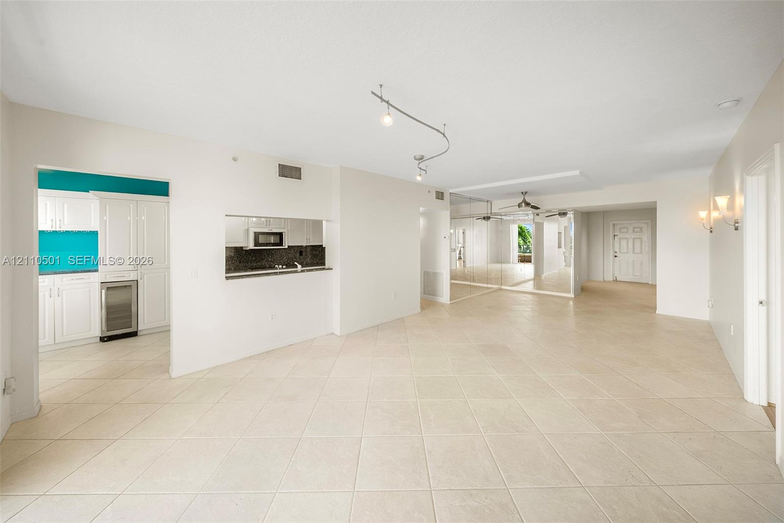 Open-concept interior featuring neutral tile flooring, track lighting, mirrored wall accents, and a kitchen service window with dark stone countertops
