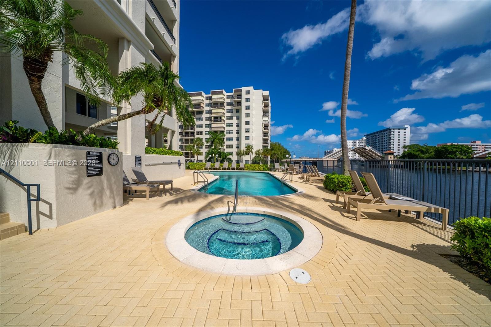 Waterfront swimming pool and circular spa featuring a herringbone-pattern paver deck, metal perimeter fencing, and mature palm landscaping