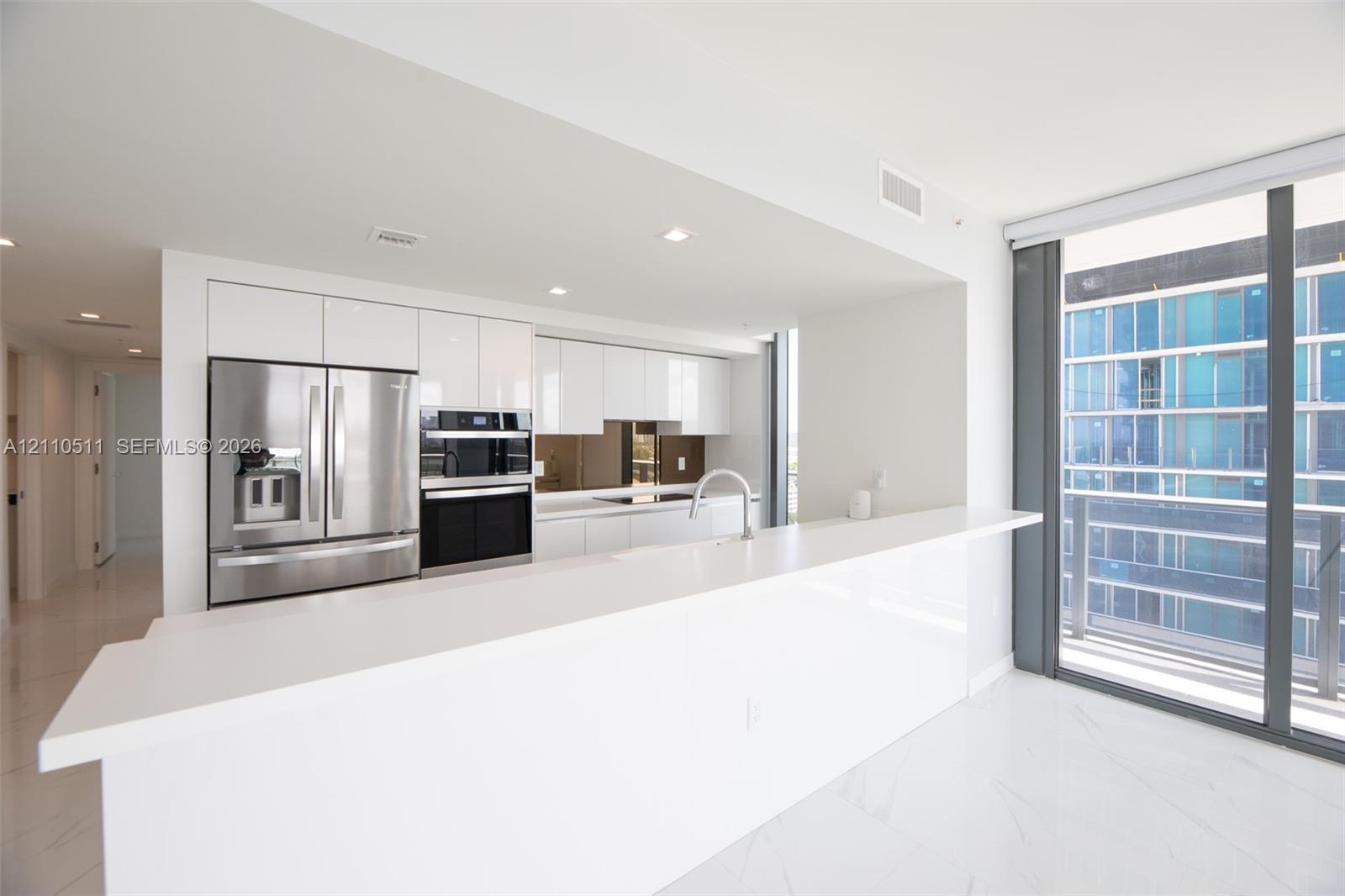 Modern kitchen featuring high-gloss white cabinetry, expansive stone countertop, stainless steel refrigerator, built-in wall oven, and floor-to-ceiling glass sliding doors