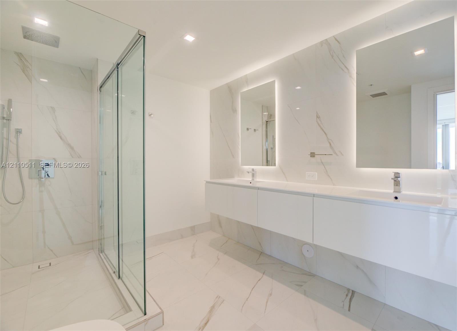 Floating dual-sink vanity with under-cabinet lighting, floor-to-ceiling stone-tiled walls, glass-enclosed walk-in shower, recessed ceiling lighting, and backlit wall mirrors