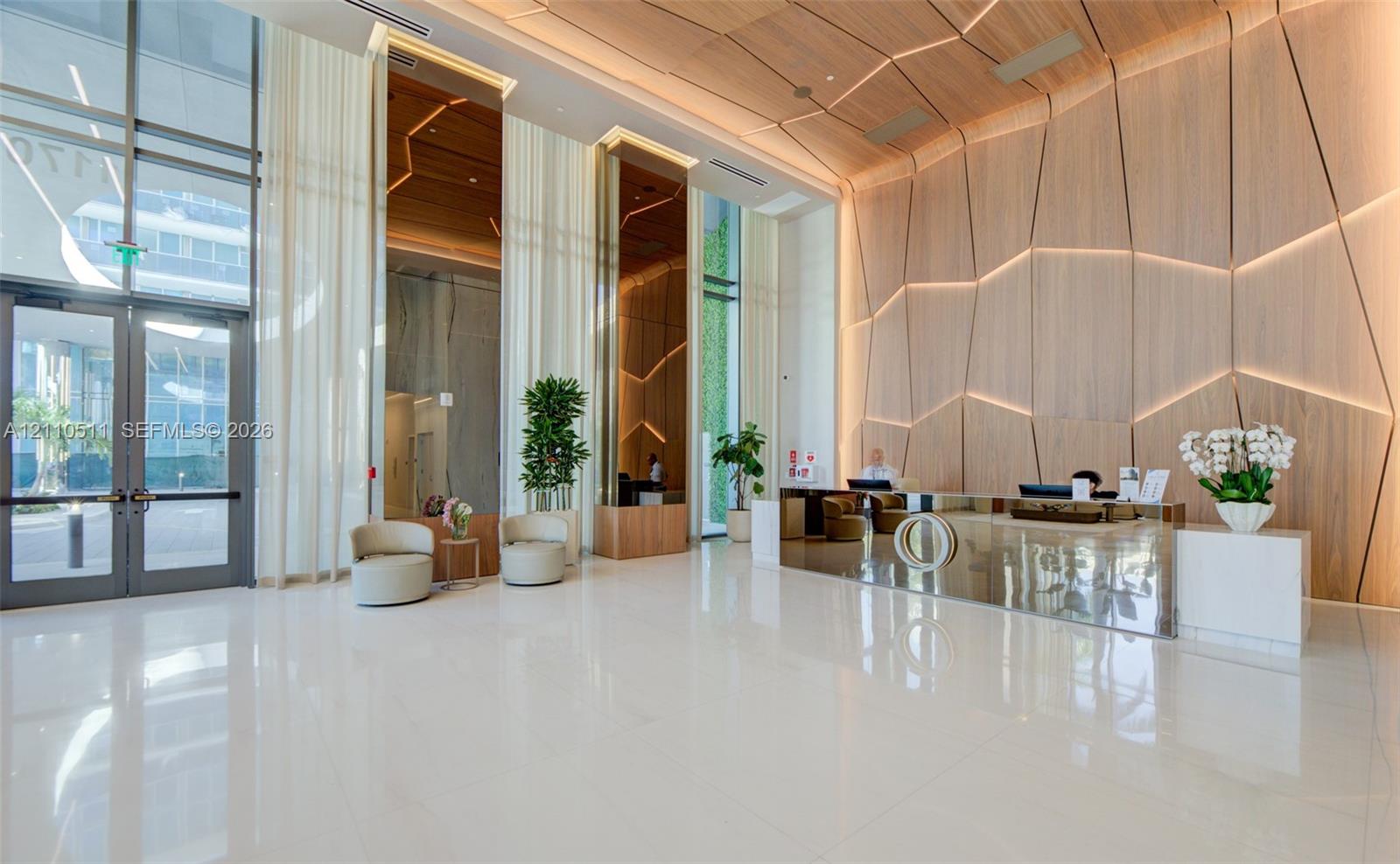 Grand lobby featuring polished white tile flooring, floor-to-ceiling glass entryway doors, geometric wood-paneled walls with integrated recessed lighting, and a mirrored reception desk