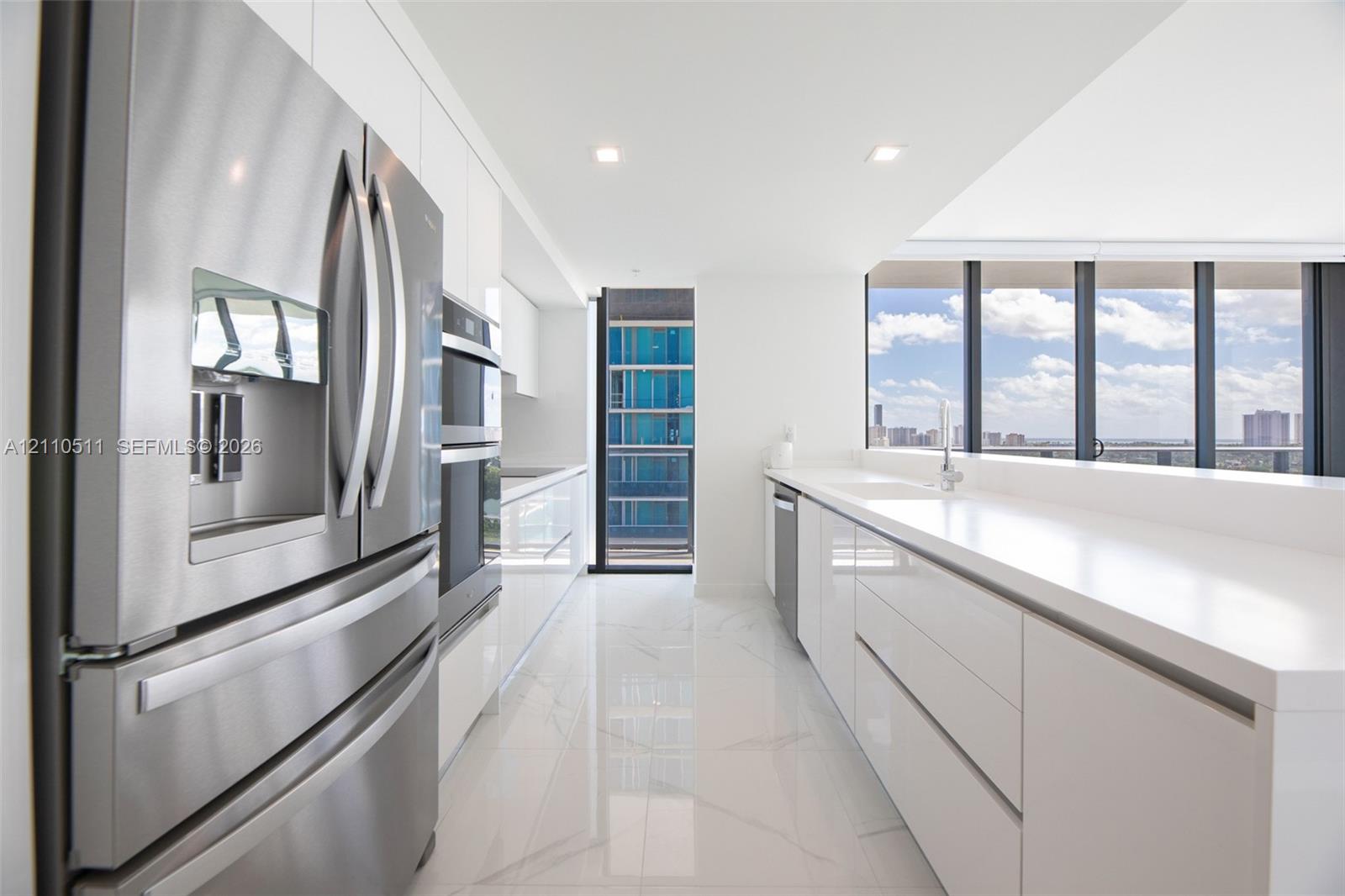 Modern kitchen featuring high-gloss white cabinetry, expansive stone countertops, stainless steel appliances, floor-to-ceiling windows, and large-format tile flooring