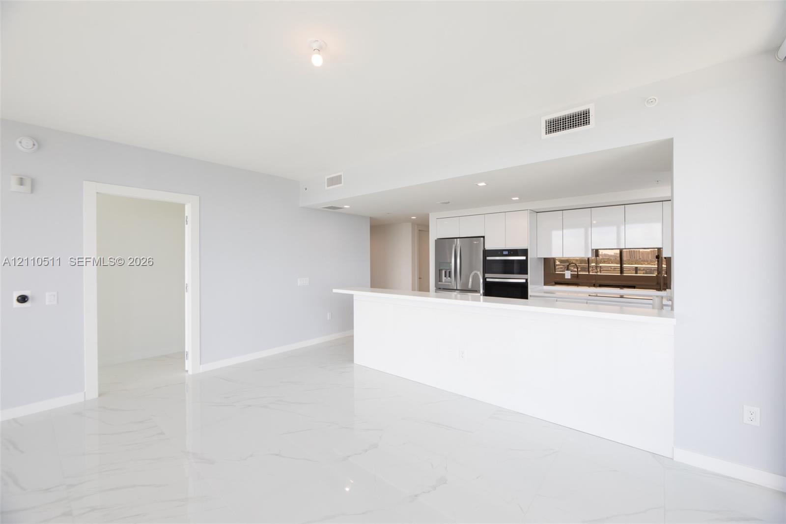 Open-concept area featuring white tile flooring with veining, white flat-panel cabinetry, a stone-surface peninsula, stainless steel appliances, and integrated recessed lighting