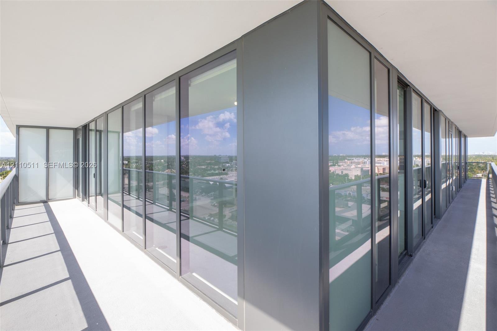 Wrap-around exterior balcony featuring floor-to-ceiling glass panels, dark metal framing, concrete flooring, and minimalist railing