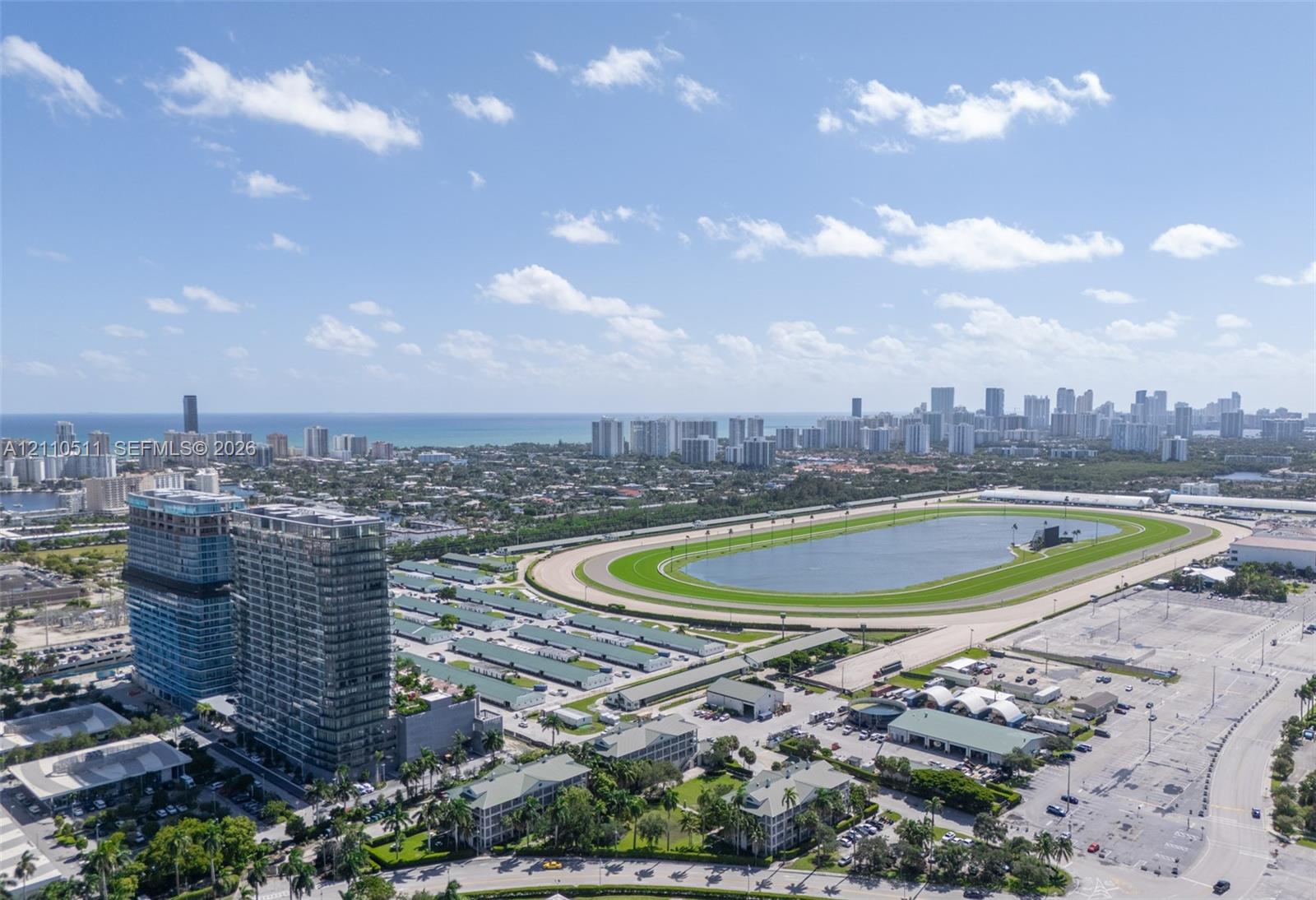 Aerial perspective featuring a multi-story glass-facade residential building, a large-scale race track, and a coastal skyline backdrop