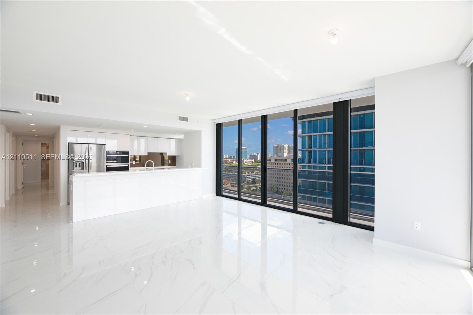Open-concept living space featuring high-gloss tile flooring, floor-to-ceiling glass windows, and a minimalist kitchen equipped with white cabinetry and stone-surfaced countertops