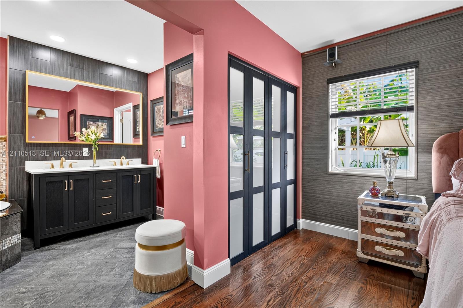 Double vanity featuring a white countertop and gold-toned hardware, large rectangular wall mirror, wood-finish flooring, dark-toned textured wall treatment, and floor-to-ceiling glass-paneled closet doors