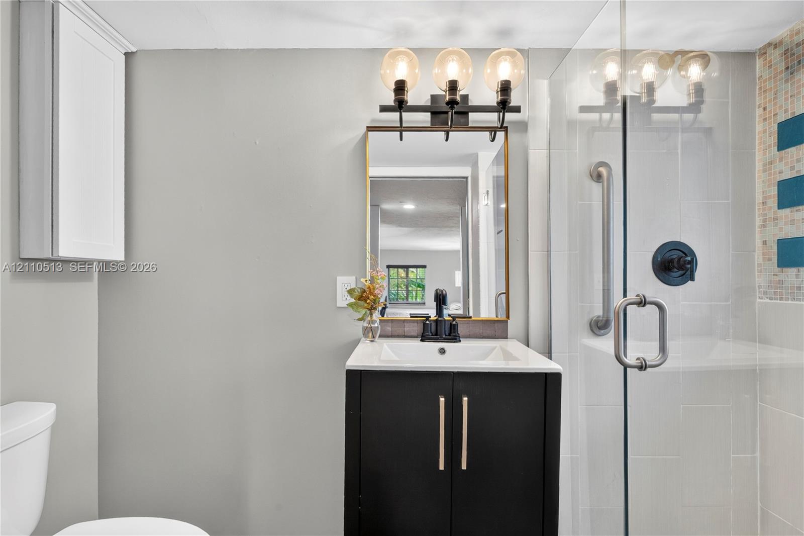 Bathroom featuring a vanity with dark cabinetry and a light-toned countertop, three-light fixture, gold-framed mirror, and a glass-enclosed shower with tiled surround